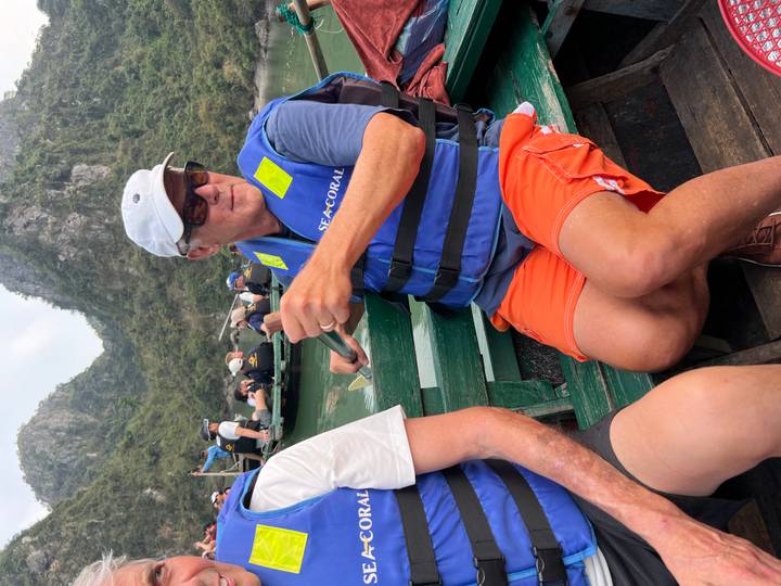 Man rows a traditional wooden skiff while fellow tourists enjoy the scenery of towering karst cliffs and calm waters.