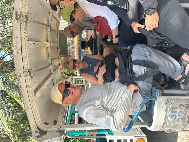 Tourists sit in the back of an open-air truck taxi under bright midday sun with palm trees overhead.