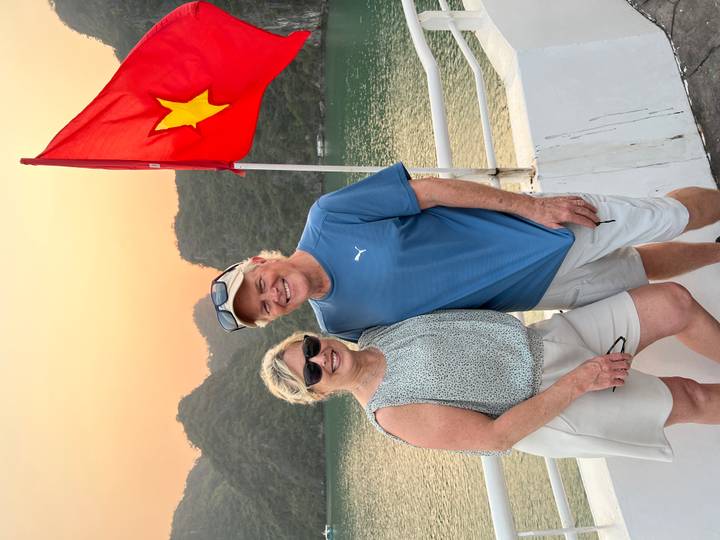 Smiling couple stands on a cruise deck at sunset with the Vietnamese flag waving and karst hills silhouetted behind.
