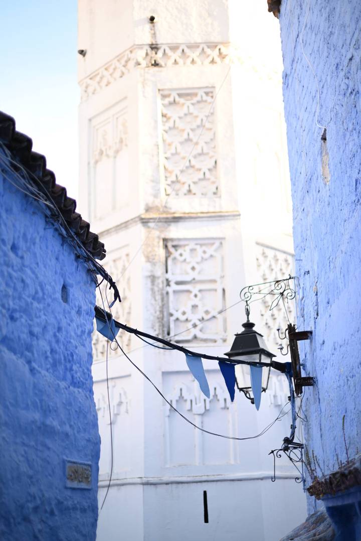 Close view of blue-washed walls and hanging wires in Chefchaouen’s medina with ornate tower blurred behind.