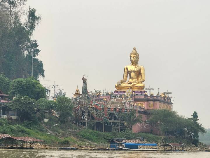 Golden seated Buddha statue atop an ornate riverside shrine in the hazy Golden Triangle region.