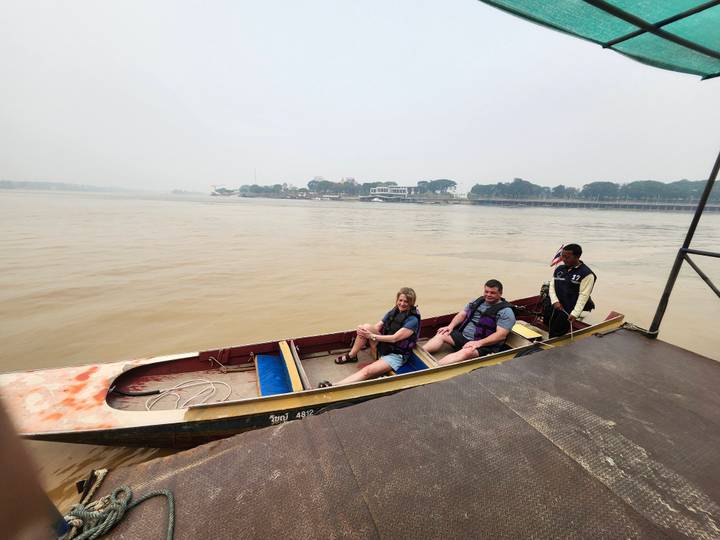 Two tourists seated in a long-tail boat with driver on a wide muddy river