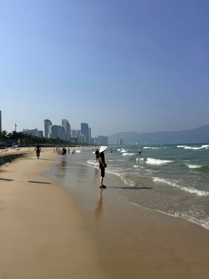 Wide sandy beach at Da Nang with city skyline and gentle waves, woman in conical hat in foreground