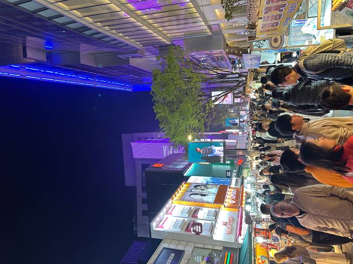 Bustling night market street packed with shoppers and neon-lit storefronts under deep blue sky.
