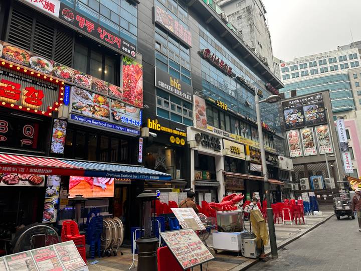 Street lined with restaurants and colourful signage in a Korean city on a gray day.