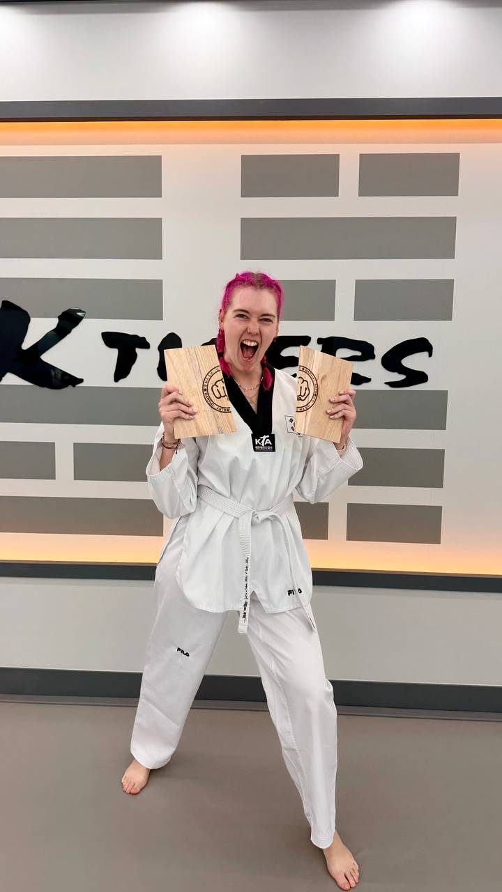 Excited woman in taekwondo uniform proudly holding two broken boards at martial arts studio.
