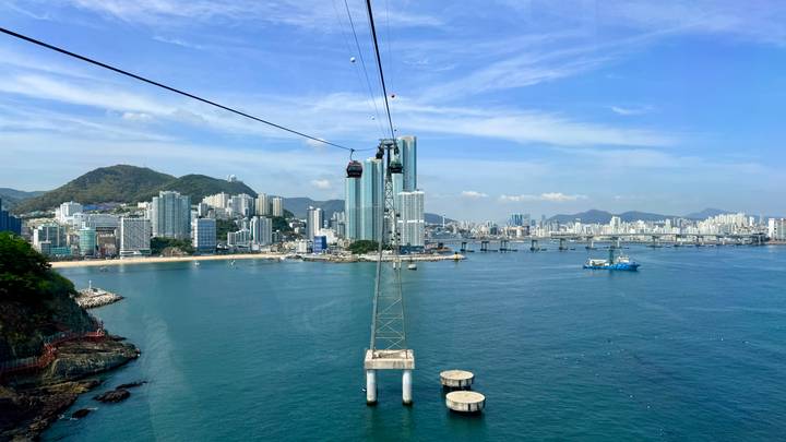 Cable car line stretching above turquoise sea toward a modern coastal skyline with surrounding hills.