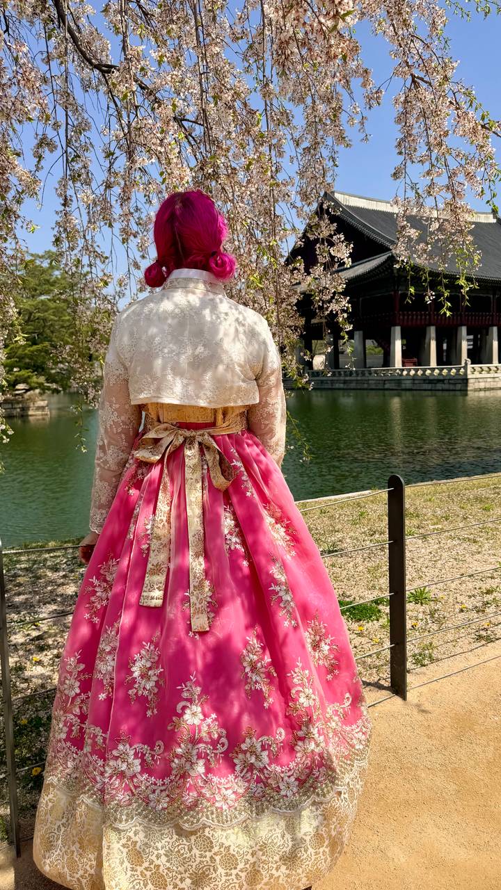 Back view of a person wearing a vibrant pink and gold hanbok standing beside a palace pond with traditional pavilion in the background.