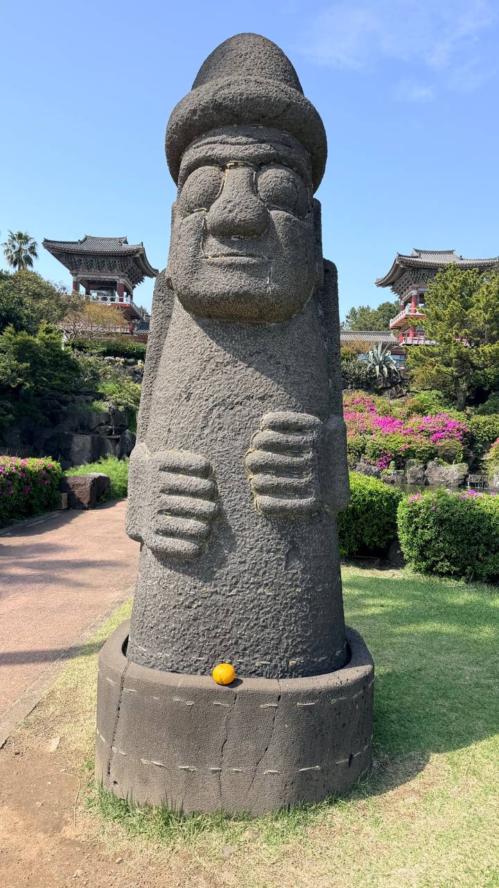 Close-up of a grey volcanic stone dolhareubang statue surrounded by blooming shrubs and a temple complex.