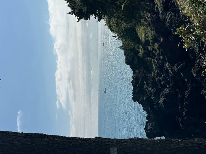 Rocky volcanic coastline overlooking a calm blue sea with sailboats on the horizon.