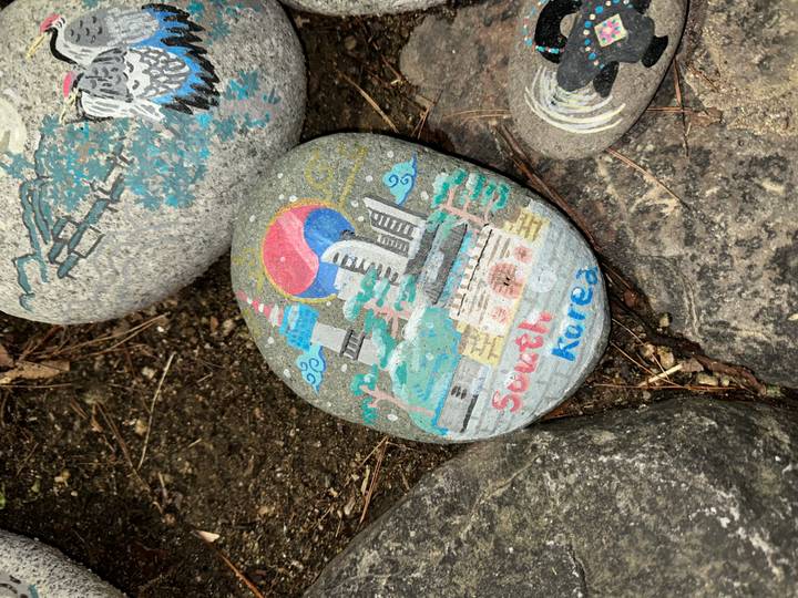 Painted souvenir stone depicting South Korea landmarks lying among rocks.