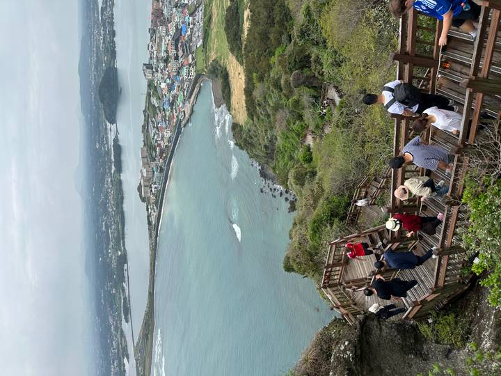 Wooden boardwalk filled with hikers descending from a volcanic peak overlooking coastal town and bay.