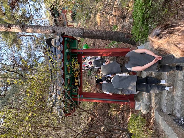 Participants in grey temple stay uniforms walking under a small wooden gate on a forested hillside.