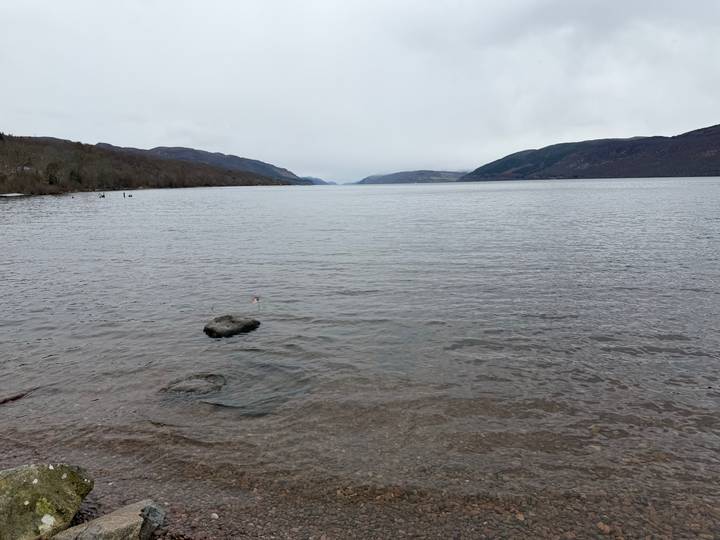 Calm, overcast view across Loch Ness with distant hills and pebbled shoreline.