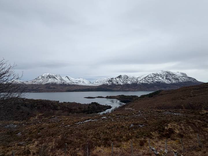 Wide view of snow-covered Torridon peaks across a still loch under grey sky.