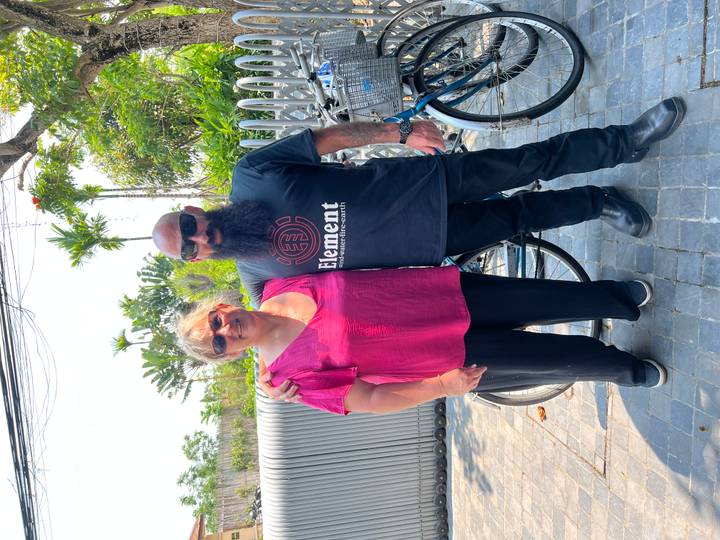 Two cyclists standing beside their bikes on a sunny paved street flanked by greenery