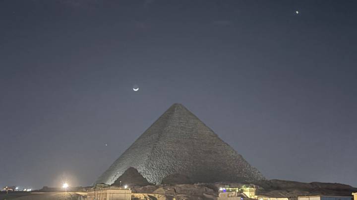 Nighttime silhouette of a pyramid at Giza beneath a crescent moon and star-dotted sky with faint ground lighting.