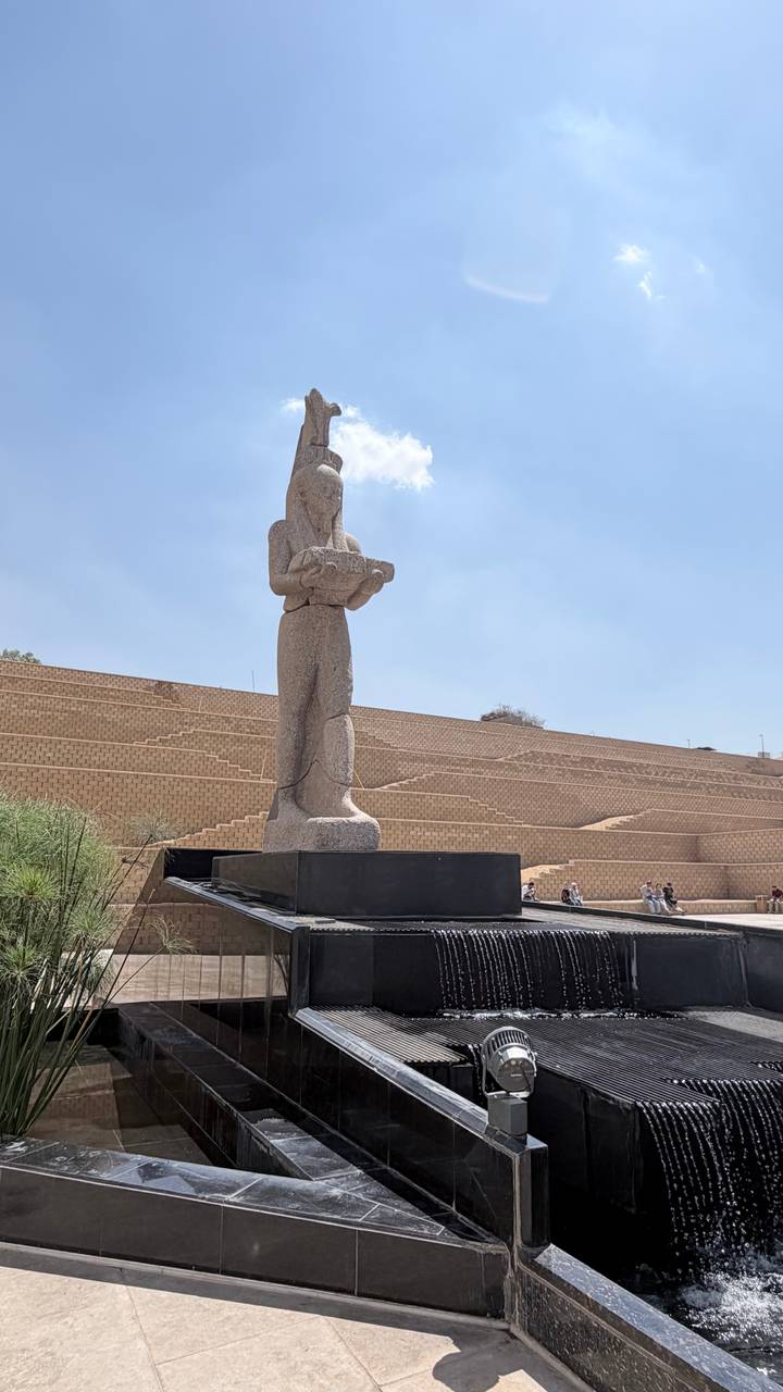 Tall stone statue of an ancient Egyptian figure displayed outdoors on a black plinth with tiered sandstone seating in the background.