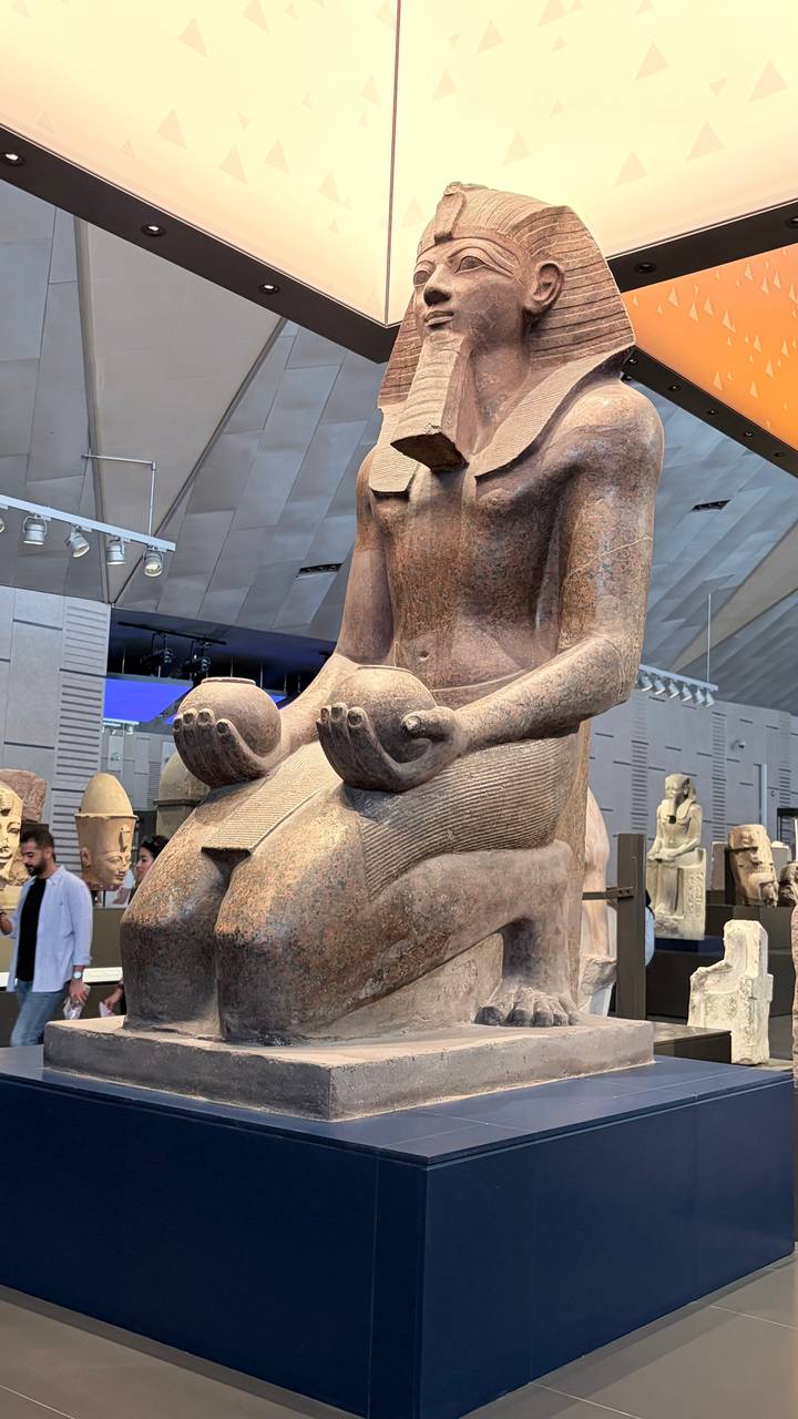 Detailed granite statue inside the Grand Egyptian Museum holding offering bowls with visitors browsing artifacts in the background.