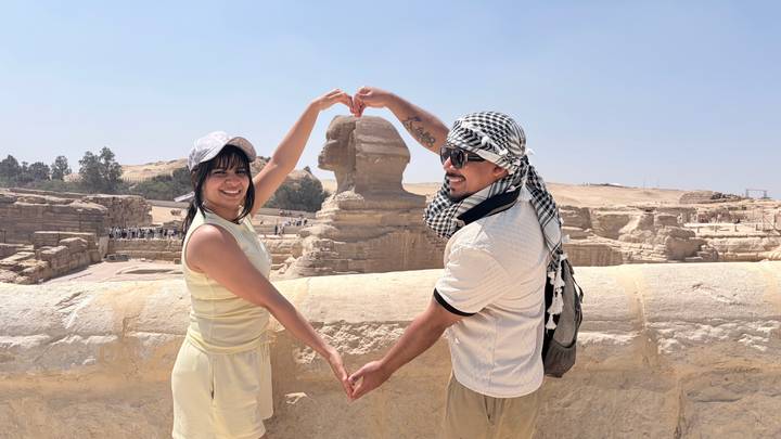 Couple makes a heart shape with their arms while posing in front of the Sphinx on the sunny Giza Plateau.