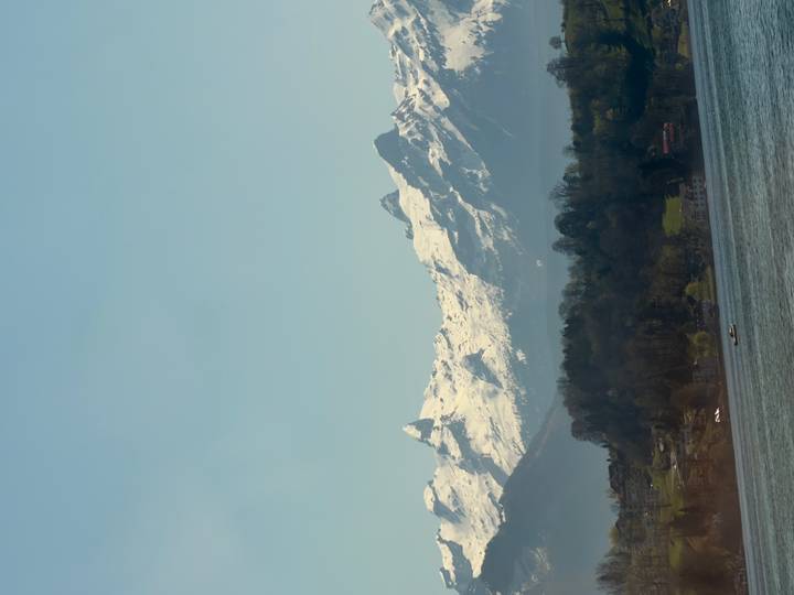 Snow-capped Swiss Alps rising above a tranquil lake with a hazy spring sky.