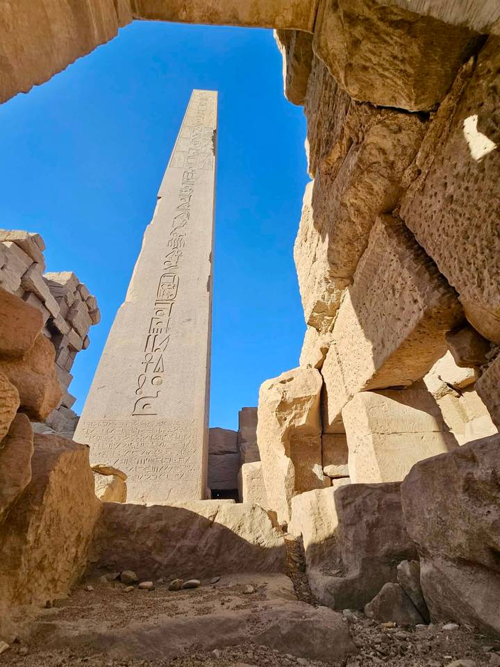Sandstone obelisk with hieroglyphs framed by massive temple blocks against clear sky.
