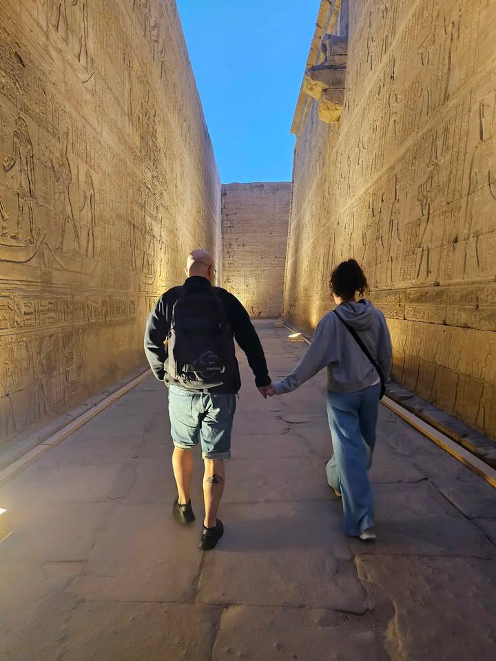 A couple walks hand-in-hand through a hieroglyph-lined corridor inside Edfu Temple.