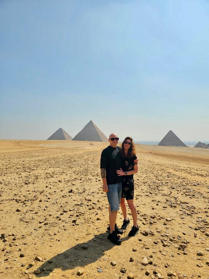 A couple embraces with the pyramids aligned in the hazy desert background.