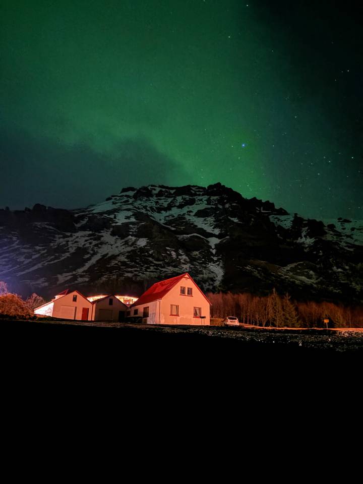 Northern lights glow green above a snow-tipped mountain and a cozy red-roofed farmhouse at night.
