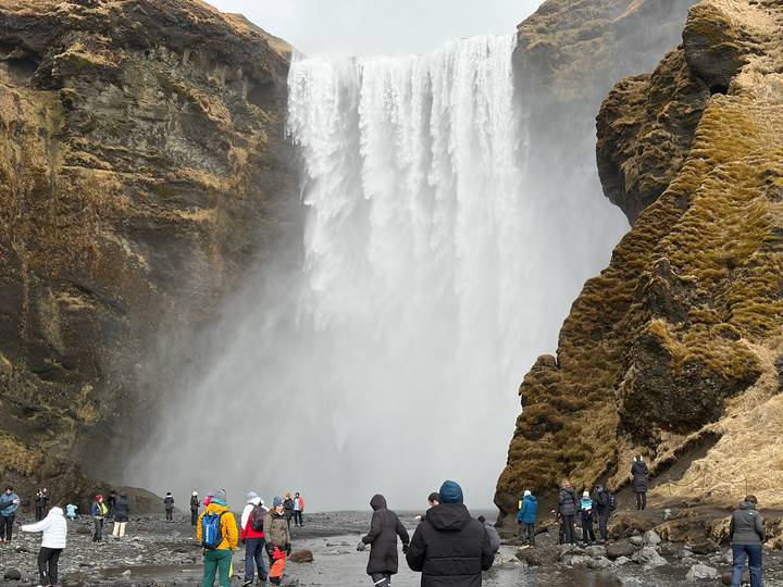 Thundering Skógafoss waterfall plunges amidst mist as bundled visitors watch from below.