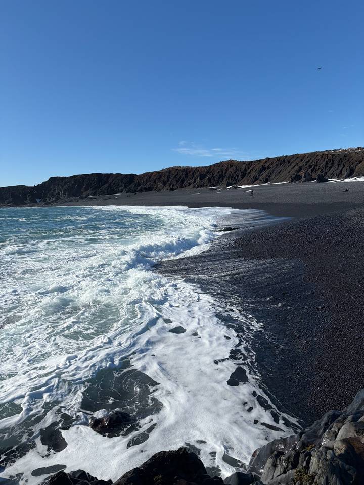 Waves rolling onto a dramatic black pebble beach with rugged cliffs in Iceland.