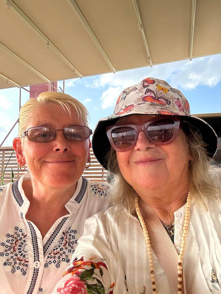 Close selfie of two women in sunglasses and sunhat smiling at an outdoor café.