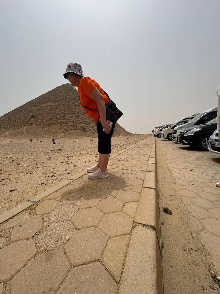 Visitor poses near the base of a desert pyramid beside a row of parked cars.