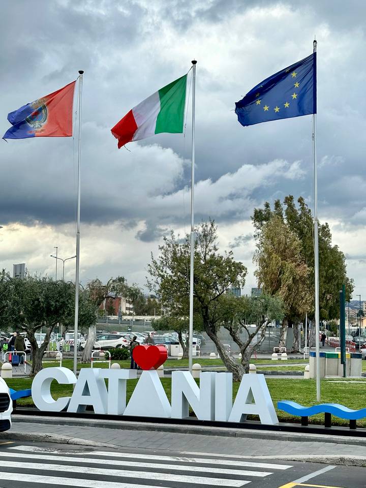 Flagpoles stand in a parking area with cloudy skies and parked cars behind.