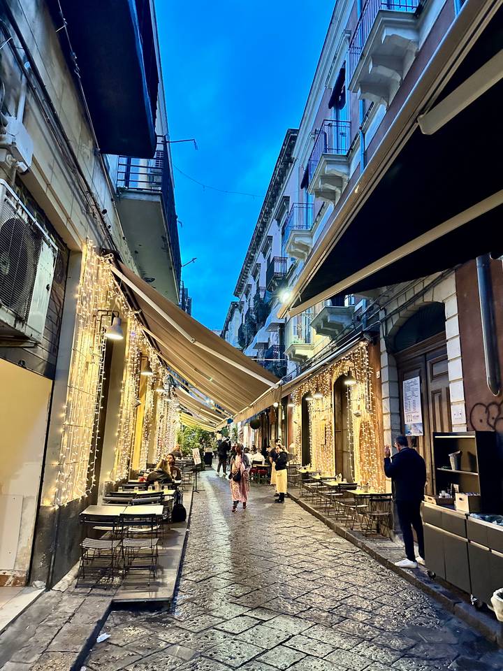 Lively narrow street lined with cafes and draped in festive string lights at dusk.