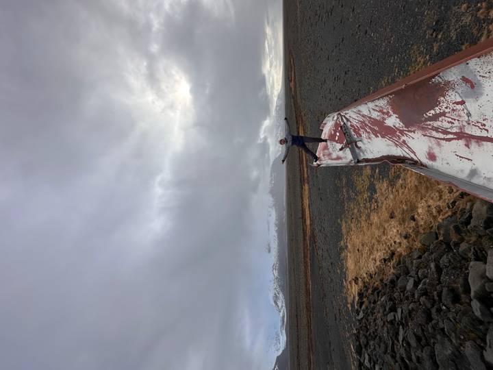 Traveller balances on the rusted hull of an abandoned vessel on a windswept Icelandic plain with snowy hills in the distance