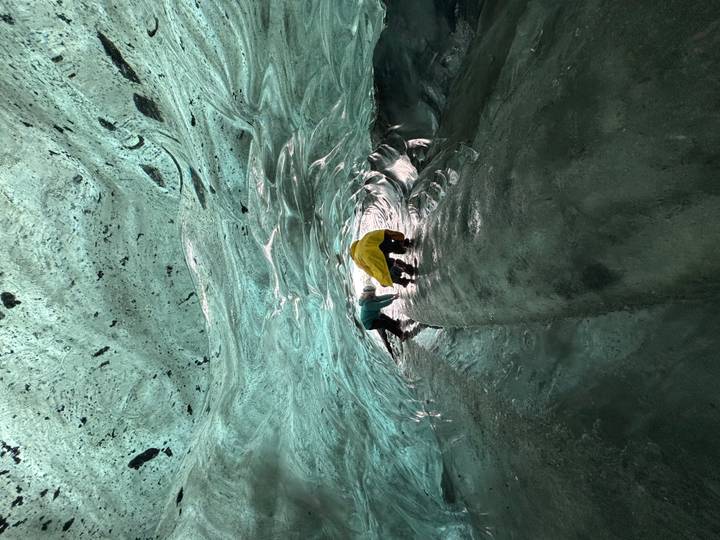 Two adventurers crawl through a shimmering turquoise ice tunnel deep inside a glacier