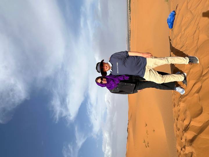Smiling couple stands atop orange Sahara dune under deep blue sky with wispy clouds.
