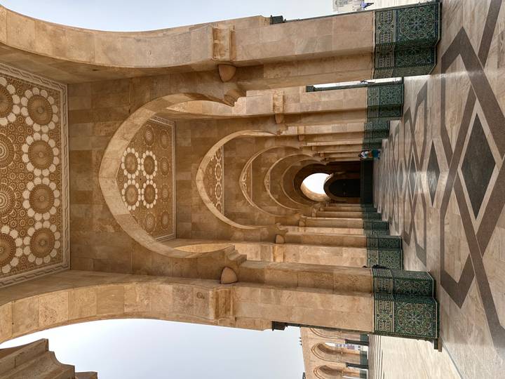 Ornate marble arches and patterned ceiling of Hassan II Mosque colonnade with a seated visitor.