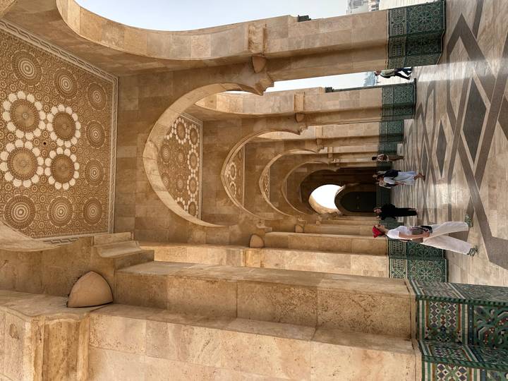 Tourists stroll through the patterned marble hallway of a grand mosque with ornate ceilings.