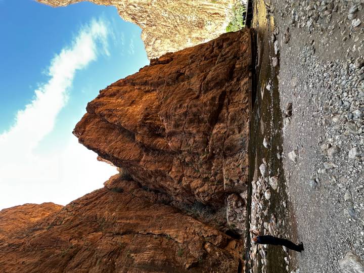 Towering orange sandstone walls of a narrow gorge with a shallow stream and rocky riverbed under a bright blue sky