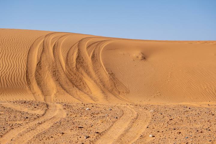 Curving tire tracks carve into smooth desert dunes under a clear pale blue sky.