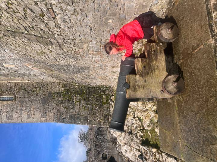 Young visitor in red jacket poses with historic brass cannon against rugged stone castle wall.