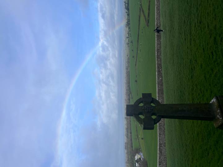 Stone Celtic cross overlooks green fields beneath a faint rainbow arching across the sky.