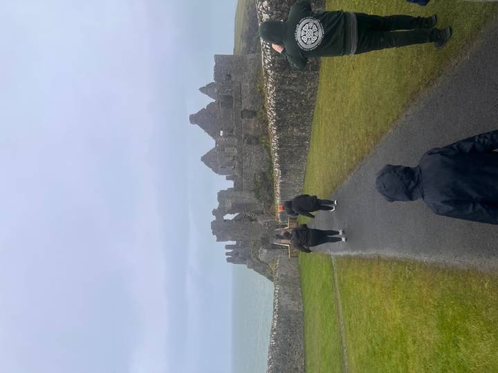 Visitors in raincoats walk toward ruins of Dunluce Castle on a windswept cliff.
