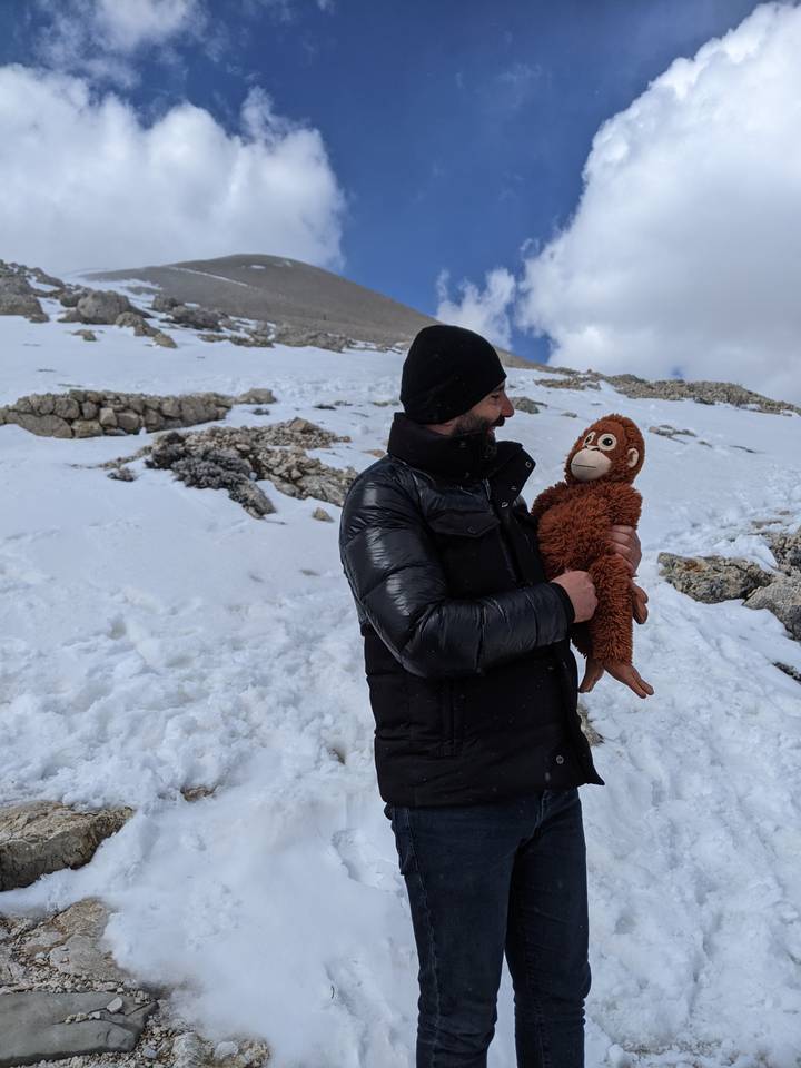 Man in a black winter coat and beanie holding a brown plush monkey while standing on a snowy mountainside.