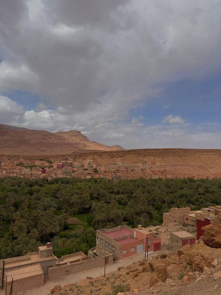 Desert valley town with red-stone cliffs rising behind a lush date-palm oasis under a partly cloudy sky.