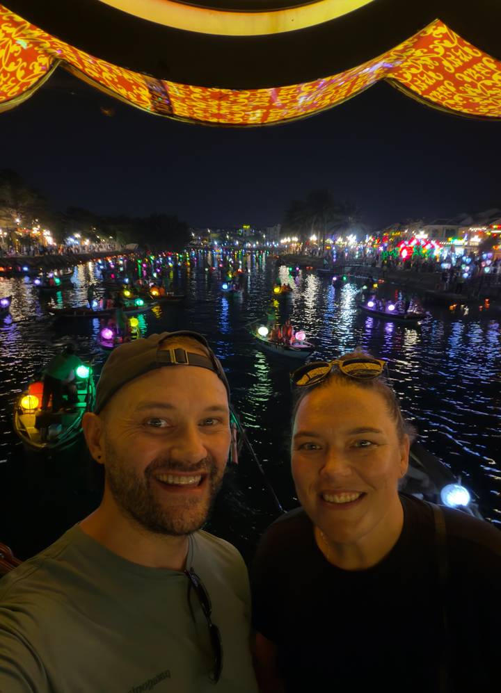 Nighttime selfie of couple with colorful lantern-lit boats floating on a river behind them