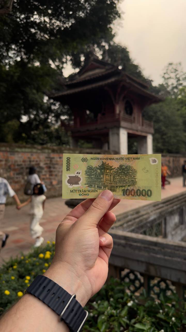 Hand holding a 100,000 VND banknote aligned with historic temple courtyard in background