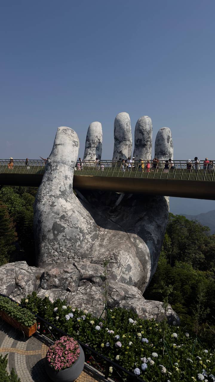 Massive stone hands support a golden pedestrian bridge high above green hills